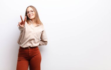 Genç ve güzel bir kadın gülümsüyor ve mutlu görünüyor, kaygısız ve pozitif, bir eliyle beyaz duvara karşı zafer ya da barış işareti yapıyor.