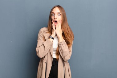 Genç ve güzel bir kadın şok olmuş ve korkmuş hissediyor. Ağzı açık ve elleri yanakları gri arka planda.