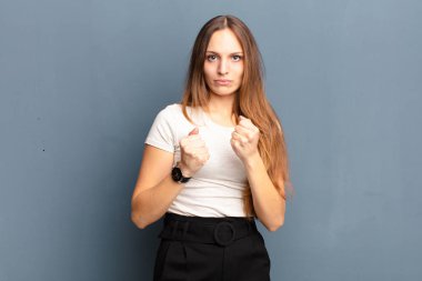 Kendine güvenen, sinirli, güçlü ve agresif görünen genç bir kadın. Yumrukları boks pozisyonunda gri arka planda dövüşmeye hazır.