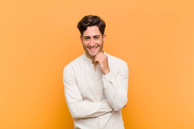 Genç yakışıklı adam mutlu görünüyor ve gülümsüyor, merak ediyor ya da soru soruyor, turuncu arka planla seçenekleri karşılaştırıyor.