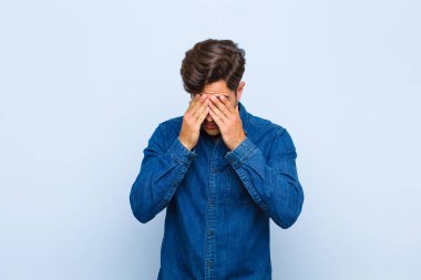 Genç yakışıklı adam üzgün, hayal kırıklığına uğramış, gergin ve depresif, iki eliyle yüzünü kapatıyor, mavi arka planda ağlıyor.