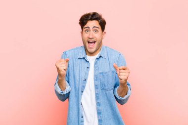 Genç yakışıklı adam çok mutlu ve şaşırmış görünüyor, başarıyı kutluyor, bağırıyor ve pembe arka plana zıplıyor.