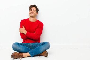 Genç yakışıklı adam kollarını kavuşturup mutlu bir şekilde gülüyor, rahat, pozitif ve tatmin olmuş bir şekilde yerde oturuyor.