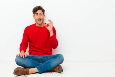 Genç yakışıklı adam kendini başarılı ve tatmin olmuş hissediyor, ağzı açık gülümsüyor, elleri yerde otururken işaret veriyor.