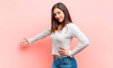 Genç, güzel bir kadın kendini mutlu ve neşeli hissediyor, gülümsüyor ve seni hoş karşılıyor, pembe arka plana karşı dostça bir jestle içeri davet ediyor..