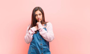 Genç ve güzel bir kadın ciddi görünüyor ve parmaklarını dudaklarına bastırıp sessizlik ya da sessizlik talep ediyor. Pembe arka plana karşı bir sır saklıyor..