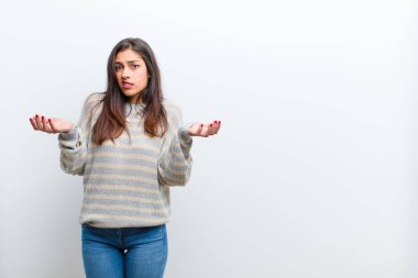 Genç, güzel bir kadın kendini cahil ve kafası karışık hissediyor, hangi seçeneği seçeceğinden emin değil, beyaz duvara yaslanmış merak ediyor.