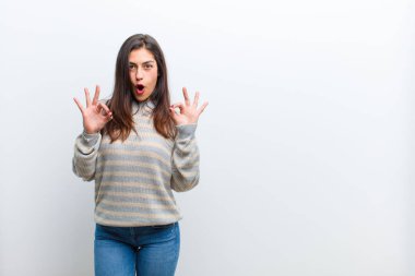 Genç, güzel bir kadın şok olmuş, şaşırmış ve şaşırmış hissediyor. Onayladığını gösteriyor. İki elini de beyaz duvara yaslamış bir şekilde işaret veriyor.