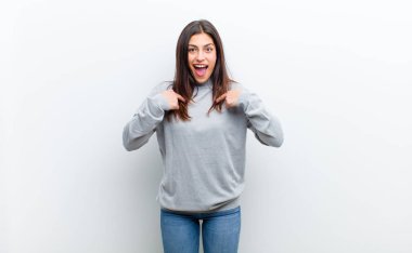 Genç, güzel bir kadın kendini mutlu, şaşırmış ve gururlu hissediyor. Heyecanlı ve şaşkın bir şekilde beyaz duvara yaslanmış gibi bakıyor.