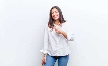 Genç, güzel bir kadın neşeyle gülümsüyor, mutlu hissediyor ve yan ve yukarı doğru işaret ediyor, kopya uzayda beyaz duvara yaslanmış nesneleri gösteriyor.