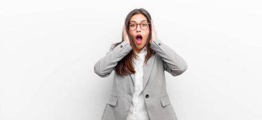 Genç ve güzel bir kadın hoş olmayan bir şekilde şok olmuş, korkmuş ya da endişeli görünüyor. Ağzı sonuna kadar açık ve iki kulağını da beyaz duvara yaslanmış ellerle kapatıyor.