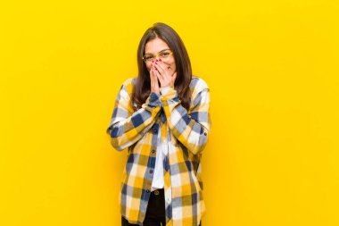 Genç ve güzel bir kadın mutlu, neşeli, şanslı ve şaşkın görünüyor. İki eli turuncu duvara dayalı ağzını kapatıyor.