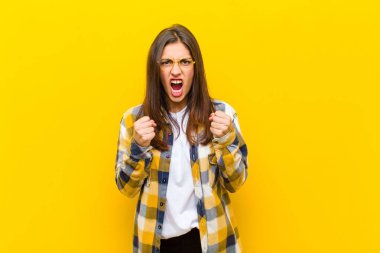 Genç ve güzel bir kadın sinirli sinirli, sinirli bakışlar ve yumruklarla bağırıyor. Turuncu duvara karşı öfkeli hissediyor.