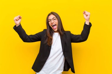 Genç, güzel bir kadın muzaffer bir şekilde bağırıyor. Heyecanlı, mutlu ve şaşırmış görünüyor. Turuncu duvara karşı kutlama yapıyor.