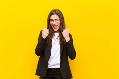 Genç ve güzel bir kadın muzaffer bir şekilde bağırıyor, gülüyor ve turuncu duvara karşı başarıyı kutlarken mutlu ve heyecanlı hissediyor.