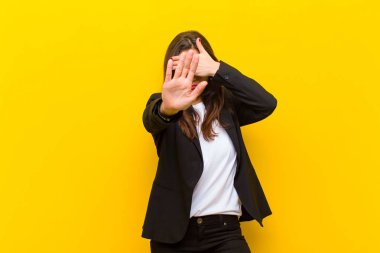 Genç ve güzel bir kadın elini yüzünü örtüyor ve kamerayı durdurmak için diğer elini öne koyuyor. Turuncu duvara karşı fotoğraf ya da resim çekmeyi reddediyor.