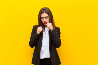 Genç ve güzel bir kadın kendinden emin, sinirli, güçlü ve agresif görünüyor. Yumrukları turuncu duvara karşı boks pozisyonunda dövüşmeye hazır.