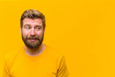 Genç sarışın adam endişeli, stresli, endişeli ve korkmuş görünüyor, panikliyor ve turuncu duvara karşı dişlerini sıkıyor.