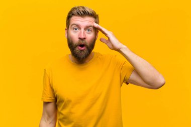 Genç sarışın adam mutlu, şaşkın ve şaşırmış görünüyor, gülümsüyor ve turuncu duvara karşı inanılmaz ve inanılmaz iyi haberler veriyor.