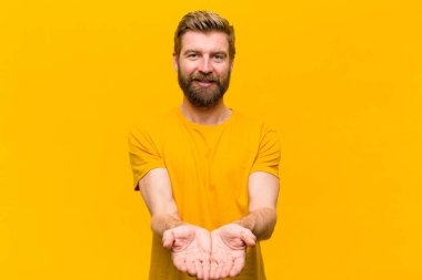 Genç sarışın adam mutlu bir şekilde gülümsüyor, kendine güveniyor, olumlu bakıyor, turuncu duvara karşı bir nesne ya da konsept sunuyor ve gösteriyor.