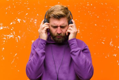 Genç sarışın adam dans ediyor ve müzik dinliyor. Kulaklıkları mor kapüşonlu. Zarar görmüş turuncu duvara karşı.