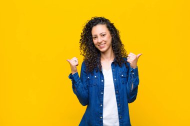 Genç ve güzel bir kadın neşeyle gülümsüyor ve mutlu görünüyor, kaygısız ve pozitif hissediyor, iki başparmağı turuncu duvara dayalı.