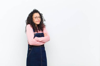 Genç güzel bir kadın omuz silkiyor, kafası karışık ve kararsız, kolları çapraz ve şaşkın bir şekilde beyaz duvara bakıyor.