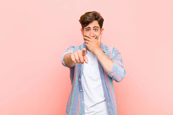 young handsome man laughing at you, pointing to camera and making fun of or mocking you against pink background