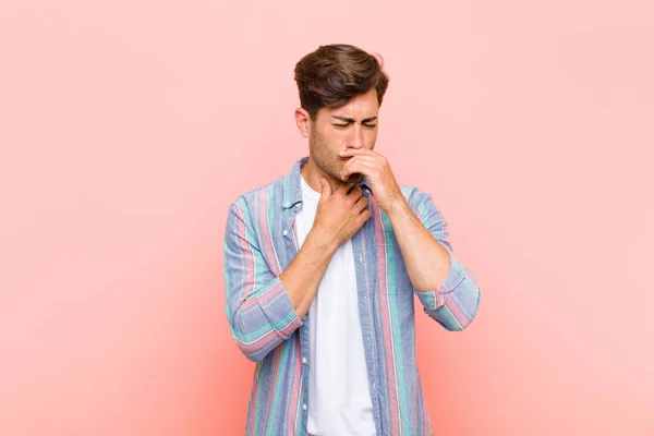 young handsome man feeling ill with a sore throat and flu symptoms, coughing with mouth covered against pink background