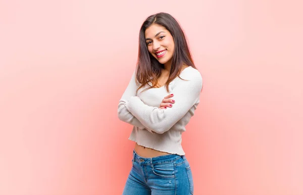 young  pretty woman smiling to camera with crossed arms and a happy, confident, satisfied expression, lateral view against pink background.