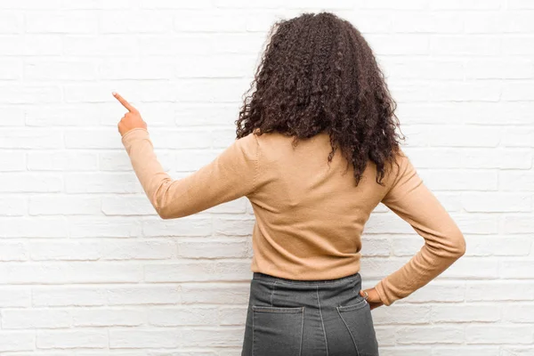 young black woman standing and pointing to object on copy space, rear view against brick wall