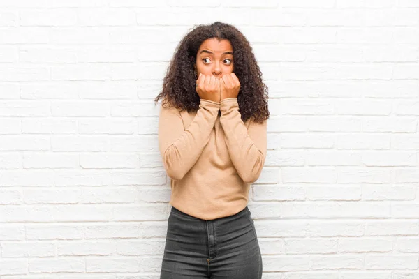 young black woman looking worried, anxious, stressed and afraid, biting fingernails and looking to lateral copy space against brick wall