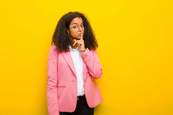 black business woman asking for silence and quiet, gesturing with finger in front of mouth, saying shh or keeping a secret against orange wall
