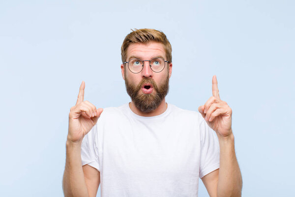 young blonde adult man feeling awed and open mouthed pointing upwards with a shocked and surprised look