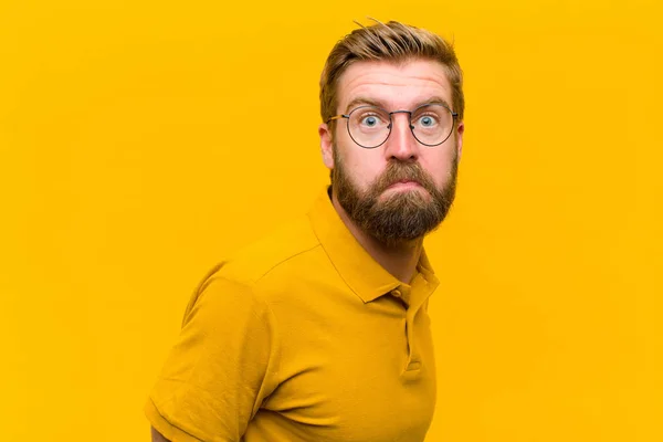 young blonde man with a goofy, crazy, surprised expression, puffing cheeks, feeling stuffed, fat and full of food against orange wall