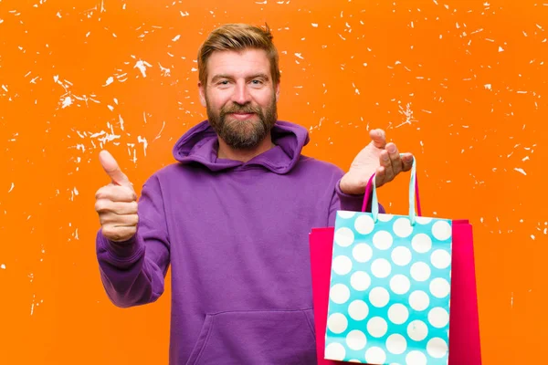 若いですブロンド男でショッピングバッグを身に着けています損傷したオレンジの壁に対して紫色のパーカー — ストック写真