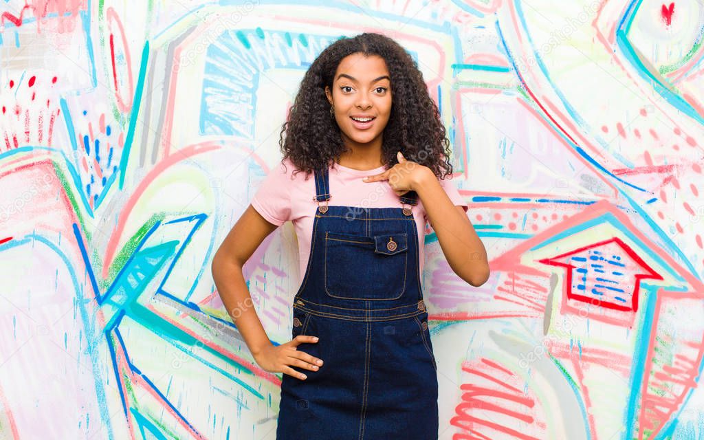 young pretty african american woman looking happy, proud and surprised, cheerfully pointing to self, feeling confident and lofty against graffiti wall