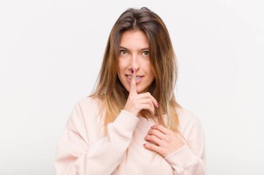 Genç ve güzel bir kadın ciddi görünüyor ve parmaklarını dudaklarına bastırıp sessizlik ya da sessizlik talep ediyor, beyaz arka plana karşı bir sır saklıyor.