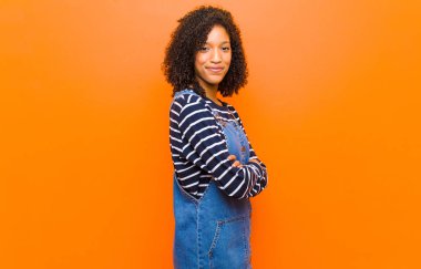 Genç ve güzel siyah kadın kameraya çapraz kollarıyla gülümsüyor ve mutlu, kendinden emin, tatmin olmuş bir ifade, turuncu duvara karşı yan bakış.