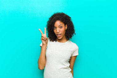 Genç siyahi kadın gülümsüyor ve arkadaş canlısı görünüyor. İki ya da iki numarayı gösteriyor. Elleri önde, mavi duvara doğru sayıyor.