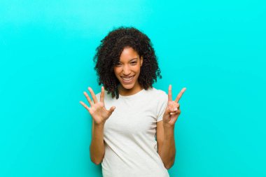 Genç siyahi kadın gülümsüyor ve dost canlısı görünüyor. Elleri önde, mavi duvara doğru geri sayıyor.