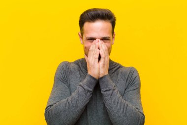 Genç yakışıklı adam mutlu, neşeli, şanslı ve şaşırmış görünüyor. İki elini de turuncu duvara dayayıp ağzını kapatmış.