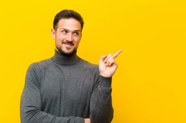 Genç yakışıklı adam mutlu bir şekilde gülümsüyor ve yan bakıyor, merak ediyor, düşünüyor ya da turuncu duvara karşı bir fikri var.