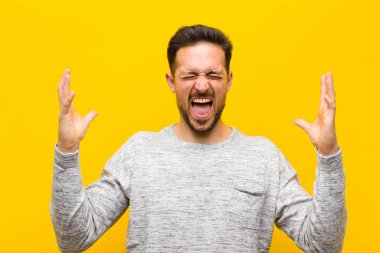 Genç yakışıklı adam öfkeyle bağırıyor, stresli hissediyor ve ellerini havaya kaldırıp neden benim turuncu duvara yaslandığımı söylüyor.