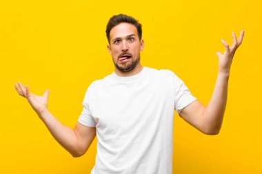 Genç yakışıklı bir adam aptal, deli, kafası karışık, şaşkın bir ifadeyle omuz silkiyor. Sinirli ve turuncu duvara karşı bilgisiz hissediyor.