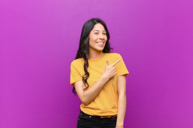 Genç, güzel, Latin bir kadın heyecanlı ve şaşırmış görünüyor. Mor duvara uzayı kopyalamak için yandan ve yukarıdan işaret ediyor.