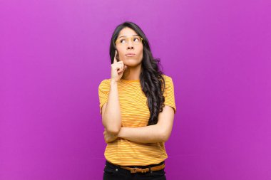 young pretty latin woman with a concentrated look, wondering with a doubtful expression, looking up and to the side against purple wall clipart