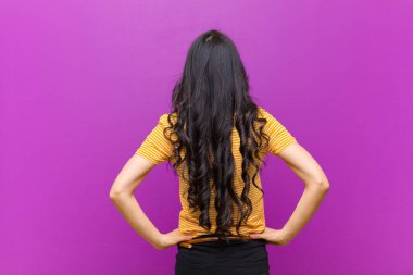 Genç, güzel, Latin bir kadın kafası karışık ya da dolu ya da şüphe ve soru dolu, merak ediyor, elleri kalçasında, mor duvarda dikiz aynasıyla