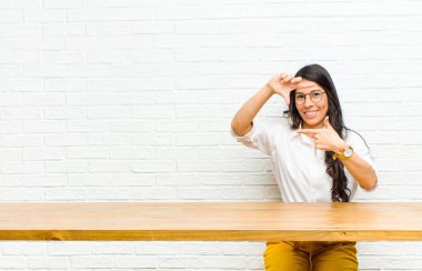 Genç, güzel Latin bir kadın mutlu, arkadaş canlısı ve pozitif hissediyor, gülümsüyor ve elleri masanın önünde oturan bir portre ya da fotoğraf çerçevesi yapıyor.