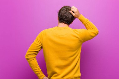 Genç, yakışıklı bir adam kendini cahil ve kafası karışık hissediyor, bir çözüm düşünüyor, eli kalçasında, diğeri kafasında, mor duvarda dikiz aynasıyla.